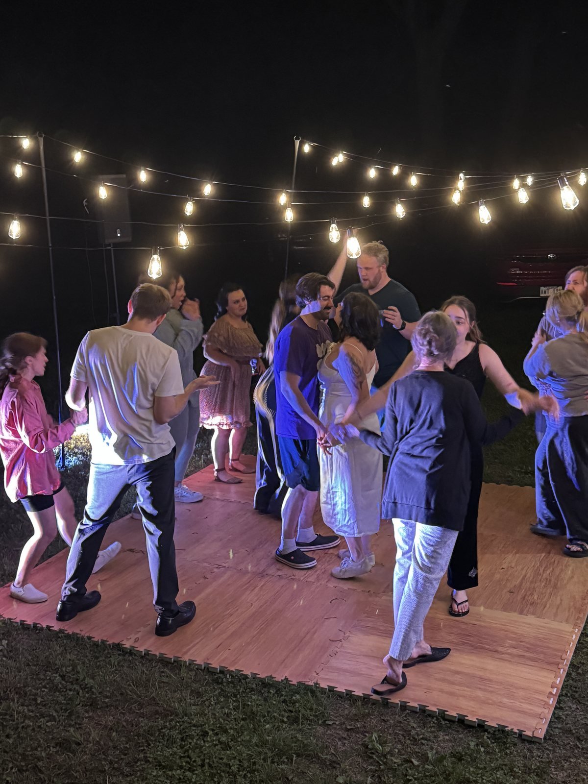 Guests dancing on an outdoor dance floor under string lights at night, with a PA speaker visible in the background